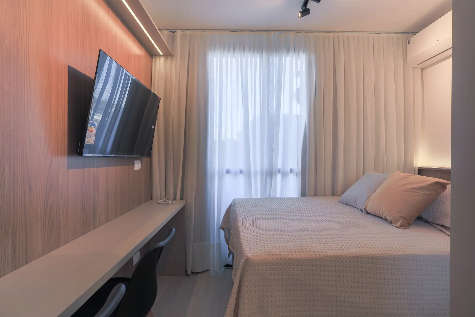 Painel de madeira com TV e bancada de trabalho, cama com almofadas e cortina de linho na janela.