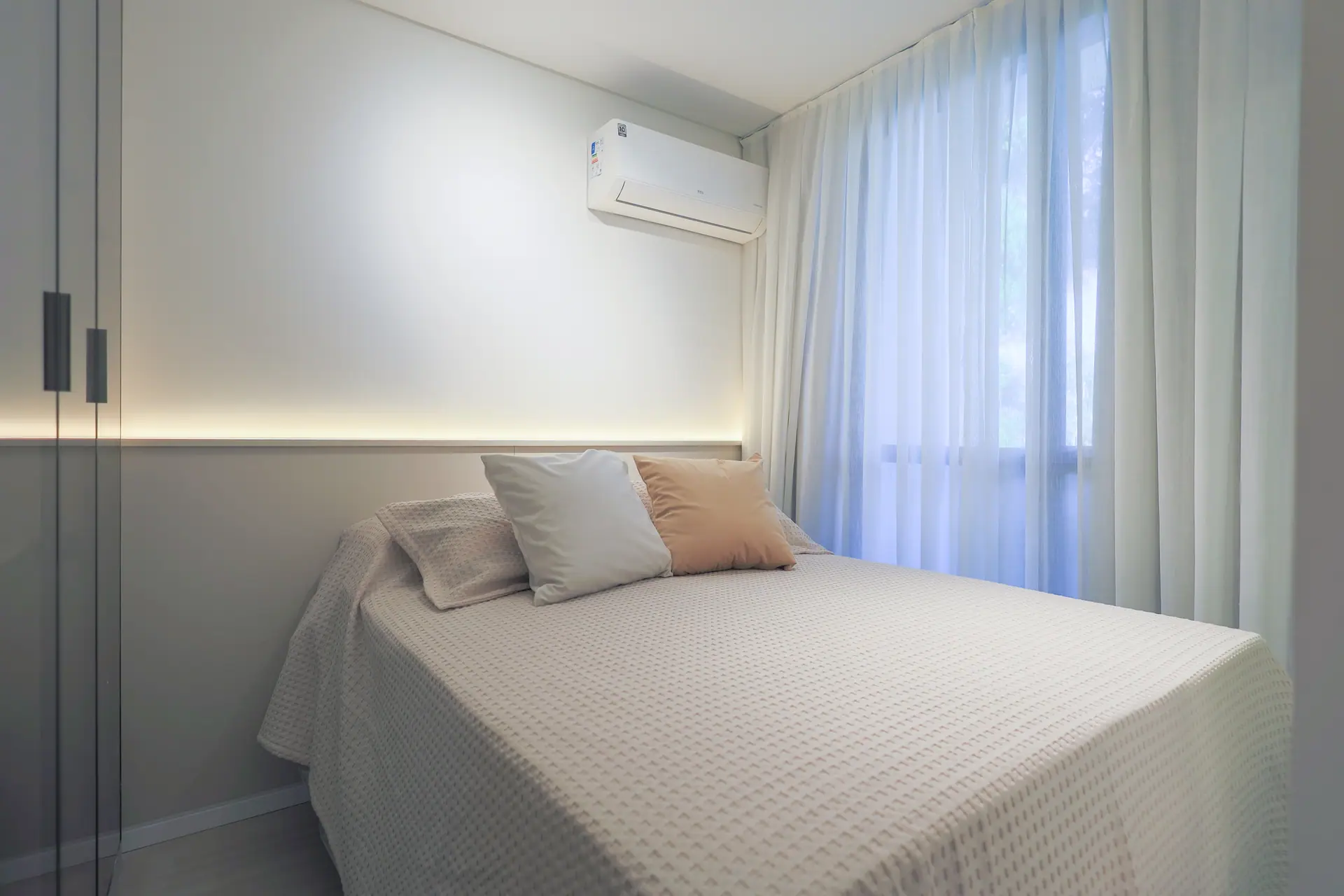 Dormitório com cama posta, luz embutida na parede e janela com cortina.